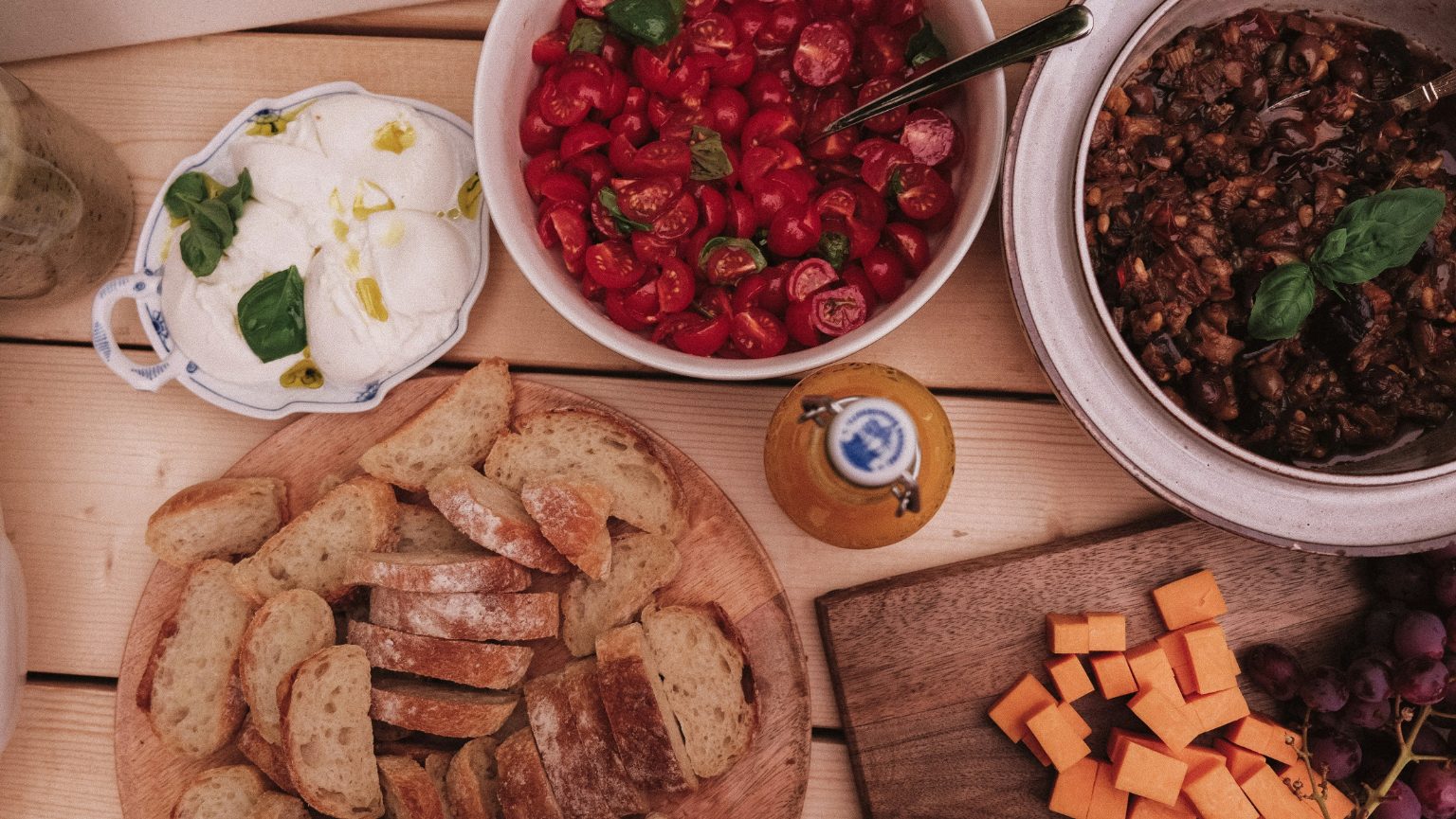 Vue du dessus d’une table en bois garnie à partager : pain tranché au milieu, burrata, tomates cerises, olives et fromage.